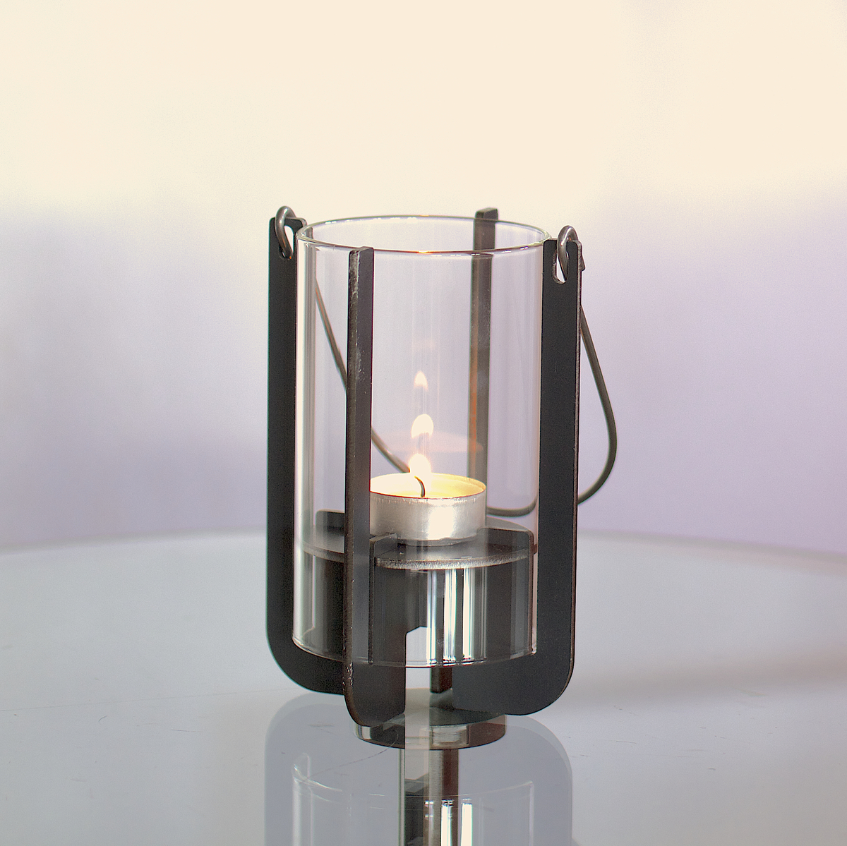 Eine moderne Gartenlaterne namens 'Lumi' steht auf einem Glas-Beistelltisch. Die Laterne ist aus schwarzem Metall und Glas gefertigt und enthält eine brennende Kerze. Im Hintergrund steht ein rostroter Sessel vor einer weißen Wand.