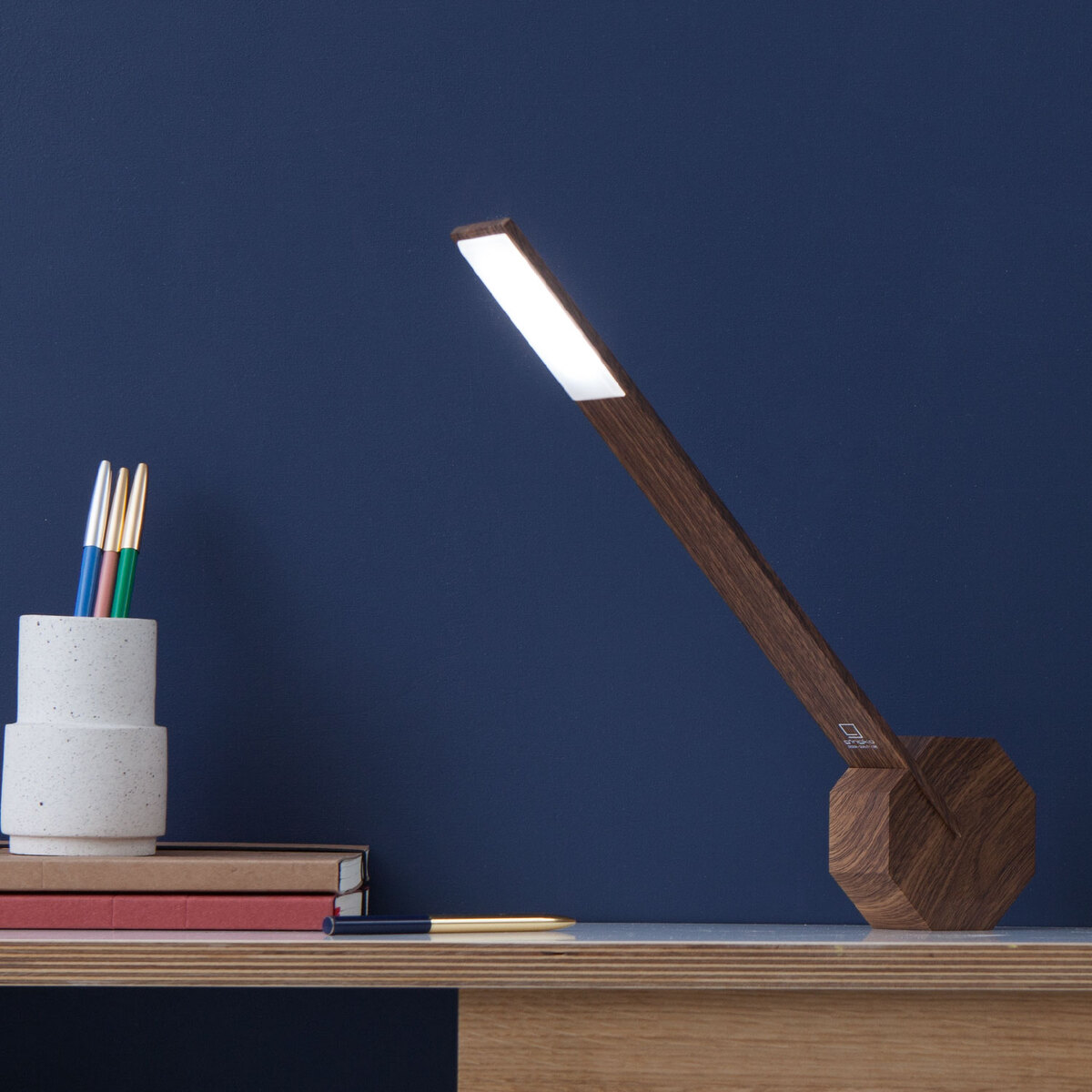 Eine moderne Tischlampe aus Holz in eindrucksvoller Octagon-Form steht auf einem Holzschreibtisch vor einer dunkelblauen Wand. Die Lampe strahlt ein helles, weißes Licht aus. Daneben stehen ein Beton-Stifteköcher mit farbigen Stiften und ein paar Bücher.