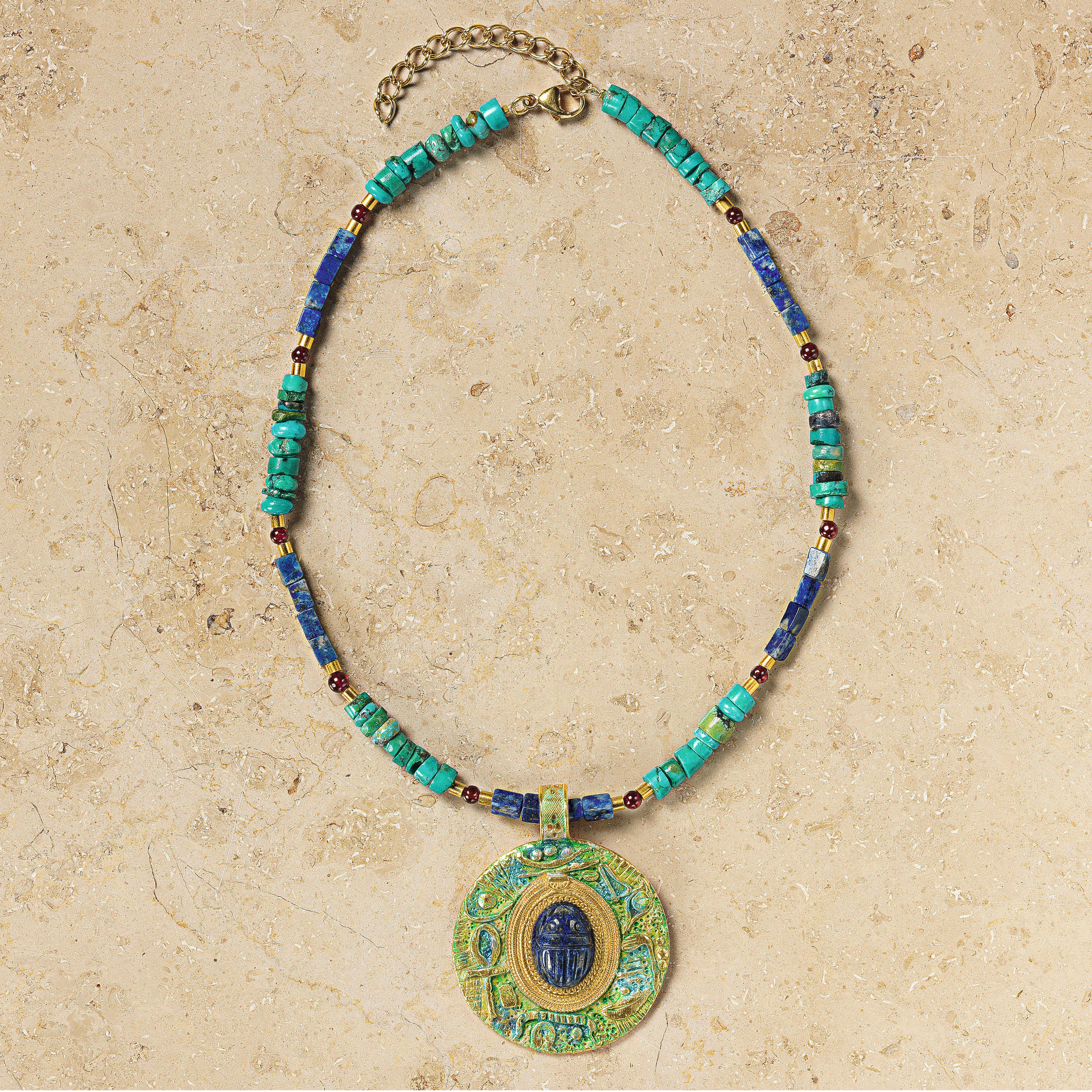 Das Collier »Scarabeo« von Petra Waszak zeigt einen zentralen Skarabäus-Anhänger aus tiefblauem Lapis in runder Form, eingefasst in vergoldetes 24-Karat-Messing. Die Kette setzt sich aus polierten Türkisen, Lapiskugeln und roten Granatperlen zusammen. Alle Elemente sind harmonisch aufeinander abgestimmt, die Oberfläche glänzt edel.