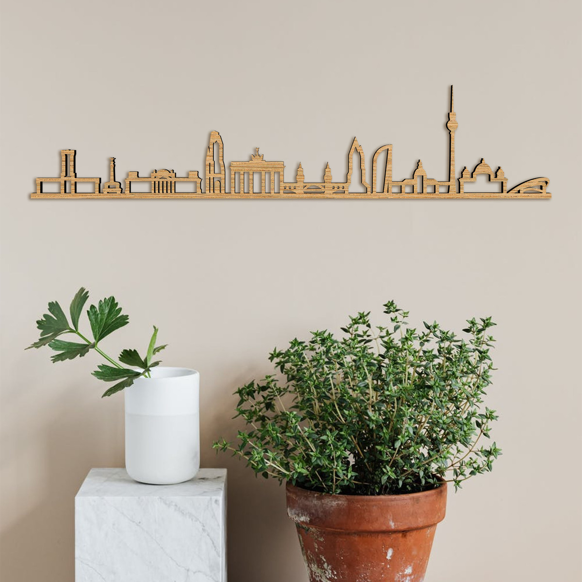 Holzkunstwerk in Form der Skyline von Berlin, aufgehängt an einer beigen Wand. Im Hintergrund sind einige Topfpflanzen und eine weiße Vase zu sehen.