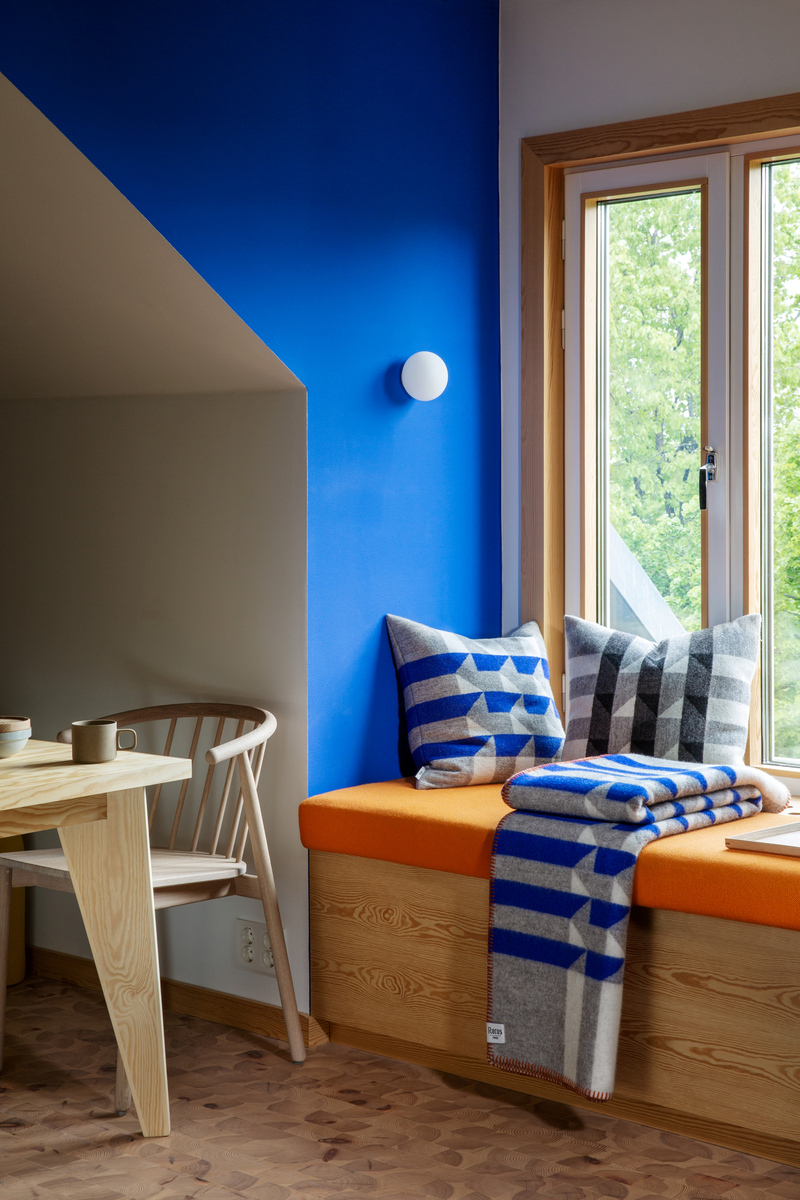Ein modernes Wohnzimmer mit einer gemütlichen Fensternische. Die Sitzbank ist mit orangefarbenen Polstern ausgestattet und mit zwei dekorativen Kissen »KVAM« in Blau und Grau mit Rautenmustern geschmückt. Ein passendes Plaid liegt auf der Sitzbank. Neben der Fensternische steht ein helles Holzmöbel-Set. Eine blaue Wand mit einer weißen Leuchte trägt zum modernen Design bei.