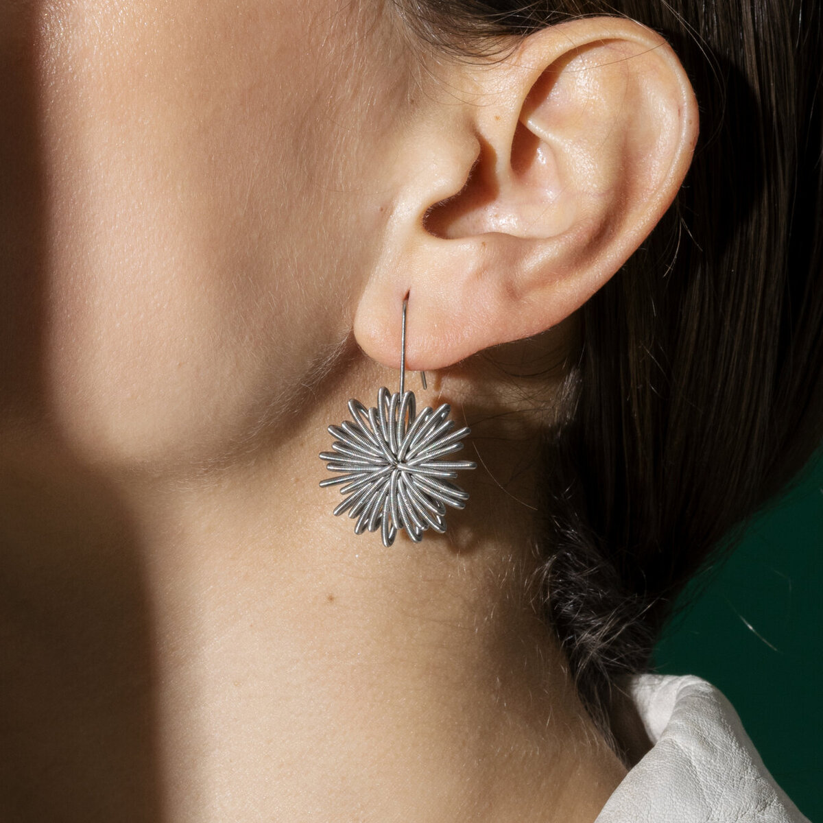 Zu sehen ist ein Ohr mit einem auffälligen, silberfarbenen Ohrring der Marke la molla. Der Ohrring „No.1 Steel“ besteht aus filigranen Edelstahlfedern in floraler Spiralform. Er sitzt leicht und elegant am Ohr. Im Hintergrund ist ein Teil eines hellen Kleidungsstücks und eine grüne Fläche erkennbar.