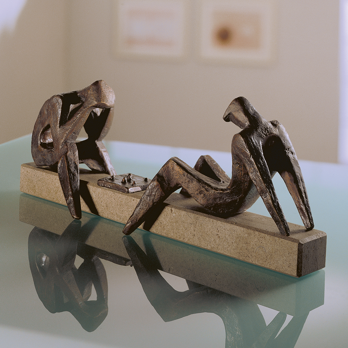 Detaillierte Bronzeskulptur von Sepp Mastaller mit zwei abstrahierten, stilisierten Personen, die Schach spielen. Die Figuren sitzen auf einem länglichen Stein-Sockel, das Schachbrett liegt zwischen ihnen. Das Kunstwerk steht auf einer reflektierenden Glasoberfläche in modernem, hellen Raum.