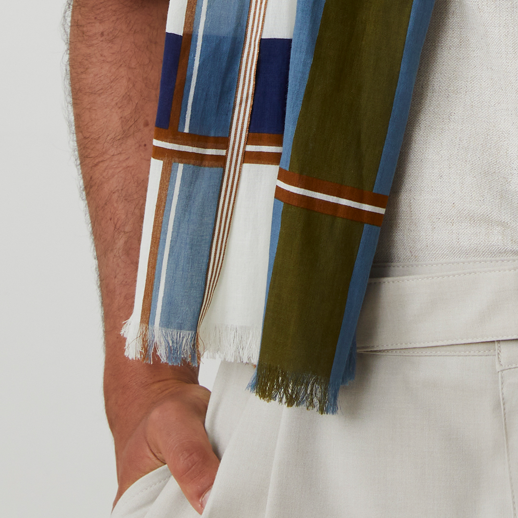 Detailansicht eines locker um den Hals gelegten Schals aus 100% Baumwolle von MOISMONT. Das Muster besteht aus breiten Längsstreifen in Blau, Grün und Weiß mit schmaleren braunen Akzentstreifen, leicht fransiger Kante und feiner Webstruktur.