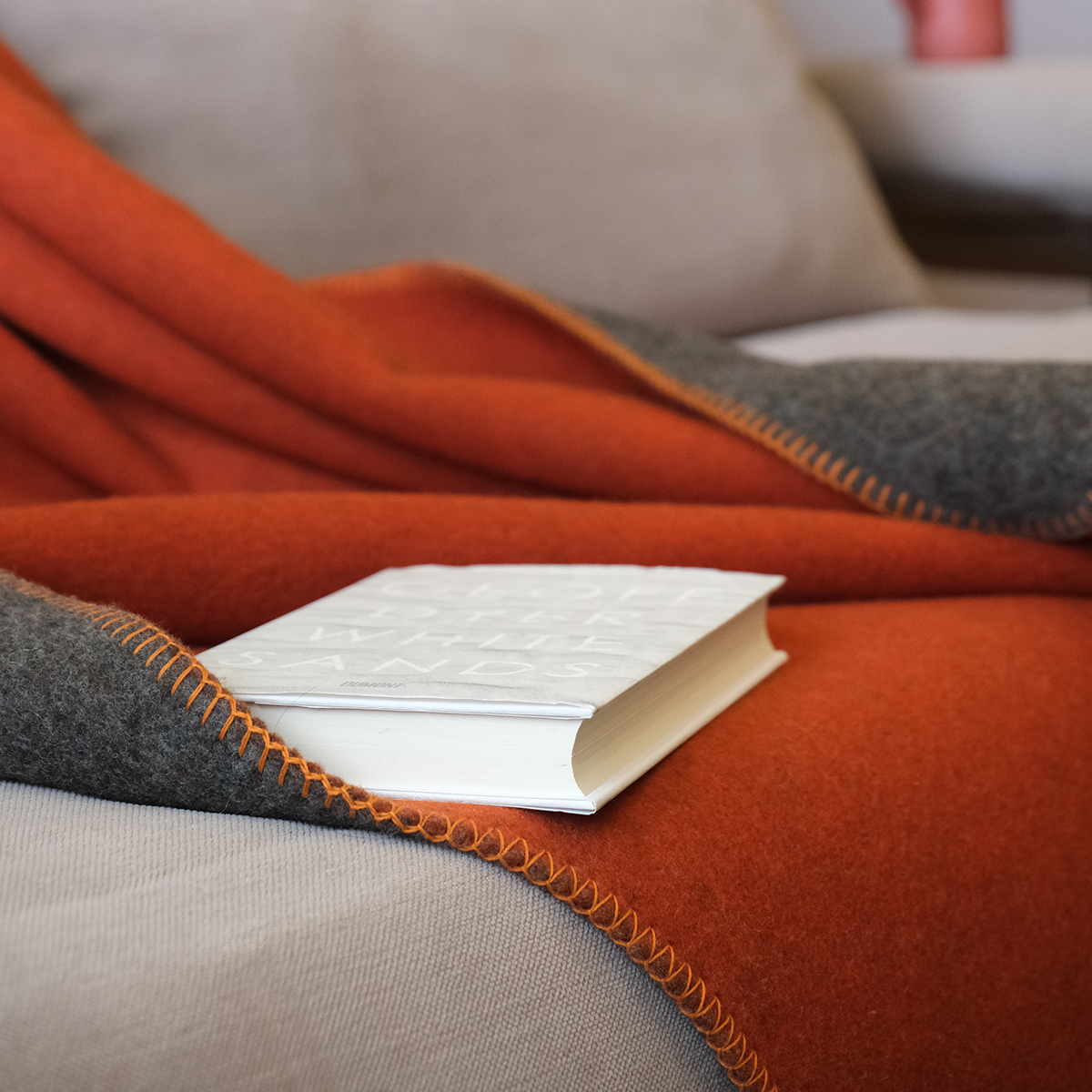 Eine Merinodecke in Orange und Dunkelgrau liegt auf einem beigen Sofa, darauf liegt ein aufgeschlagenes Buch. Die Szene wirkt gemütlich und lädt zum Entspannen und Lesen ein.