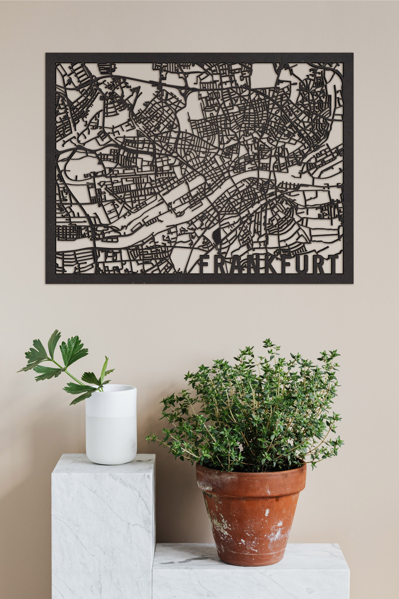 Der Stadtplan von Frankfurt aus Holz zeigt filigran die Straßen und Gebäude der Stadt. Das Kunstwerk hängt an einer beigen Wand über einem weißen, marmorähnlichen Sockel und einem Terrakottatopf mit einer grünen Pflanze.