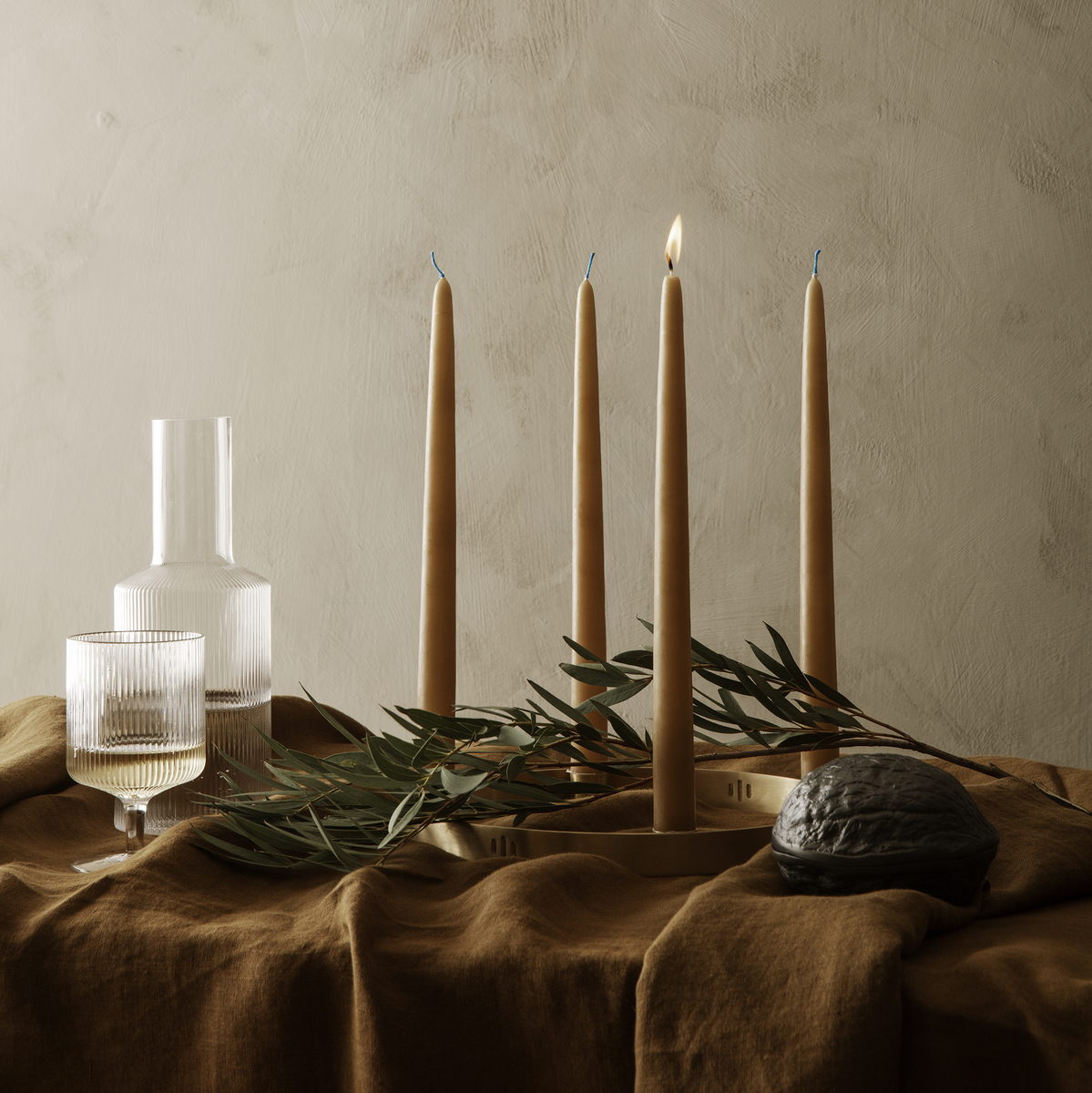 Stilvolle Tischdekoration mit einem minimalistischen Kerzenhalter 'Circle' und mehreren brennenden Stabkerzen. Der Tisch ist mit braunem Stoff bedeckt, umgeben von Olivenzweigen und verschiedenen Glasgefäßen.