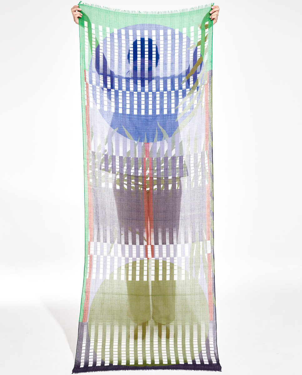 Ein Schal aus Baumwolle und Seide, mit einem geometrischen Muster aus Grün, Blau, Violett, Weiß und Rot, gehalten von einer Person vor einem hellen Hintergrund.