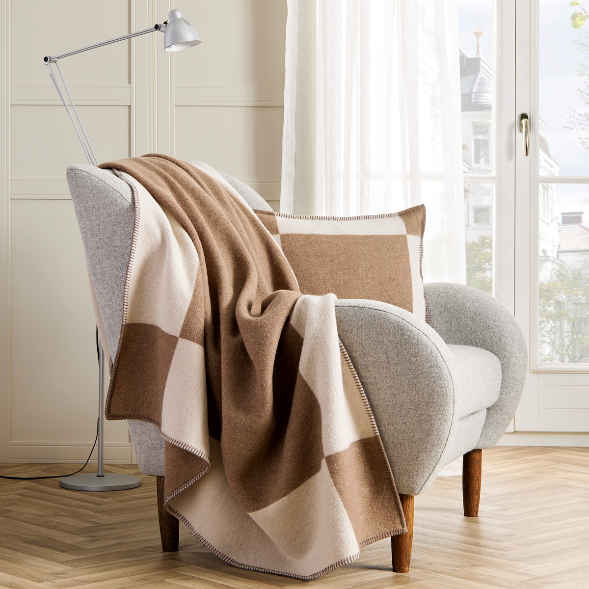 Eine gemütliche Decke »Manchester« in Beige- und Brauntönen liegt dekorativ über einem grauen Sessel in einem hellen und modernen Wohnzimmer. Im Hintergrund sind ein großes Fenster und ein heller Holzfußboden zu sehen.