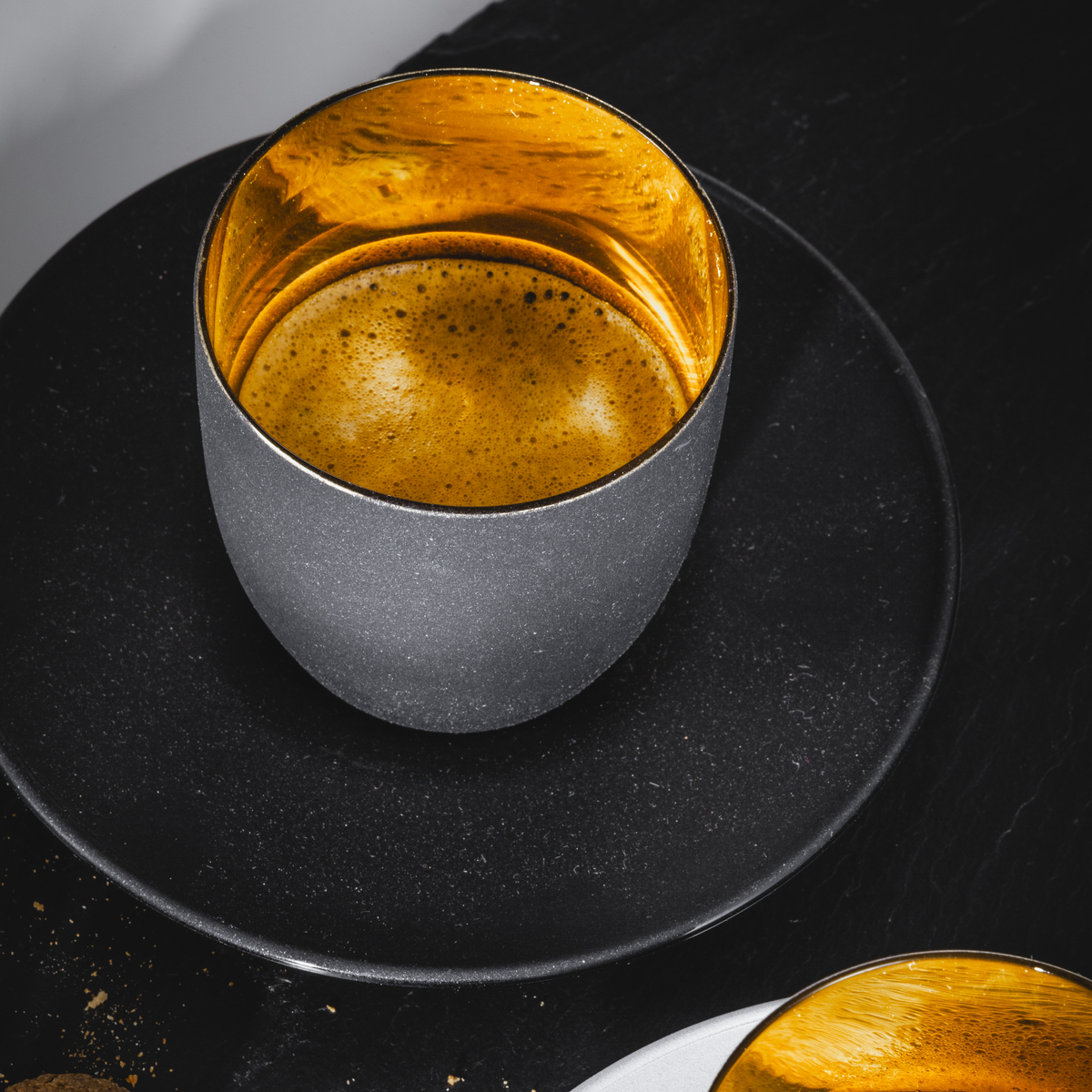 Zwei moderne Espressobecher auf schwarzen und weißen Untertassen, einer mit goldener Innenbeschichtung, gefüllt mit Espresso. Im Vordergrund befinden sich einige Keksstücke und Krümel. Das Bild ist stilvoll arrangiert und hebt die Eleganz der Kaffeetassen hervor.
