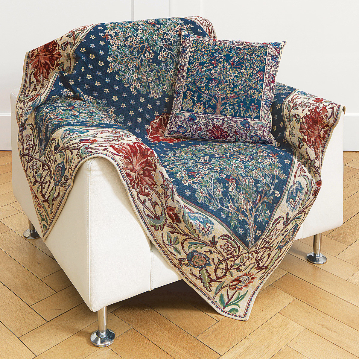 Ein weißer Sessel ist mit einer Wohnplaid und einer passenden Kissenhülle im 'Tree of Life'-Design nach William Morris geschmückt. Die Textilien zeigen ein reich verziertes florales Muster in Blau, Creme, Rot und Grüntönen. Das Set betont einen klassischen, künstlerisch inspirierten Einrichtungsstil im Wohnzimmer.