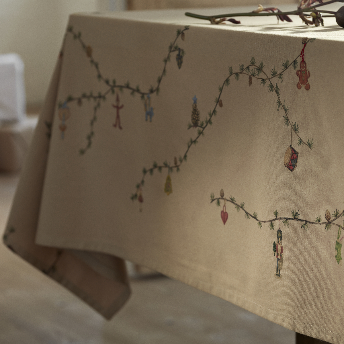 Beigefarbene Tischdecke mit gestickten grünen Zweigen und bunten Weihnachtsmotiven, dekorativ auf einem Esstisch arrangiert, sanft beleuchtet, geeignet für festliche Anlässe.