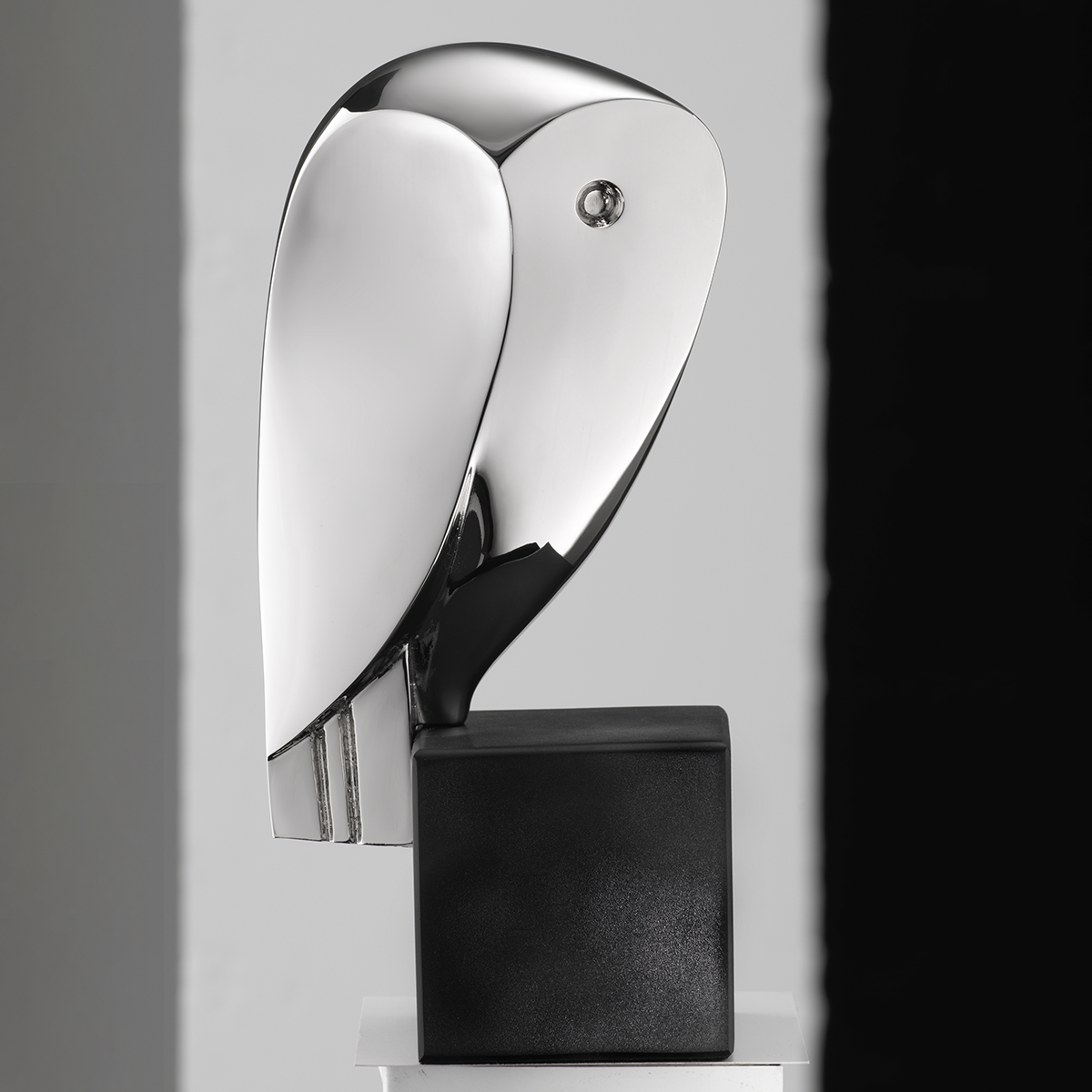 Abgebildet ist eine elegante, moderne Skulptur eines stilisierten Vogels der Künstlerin Cordula Dobes in metallic-silber auf einem schwarzen, quaderförmigen Sockel. Die Oberfläche glänzt spiegelnd. Die Skulptur steht vor einem minimalistischen, hell-dunkel geteilten Hintergrund.