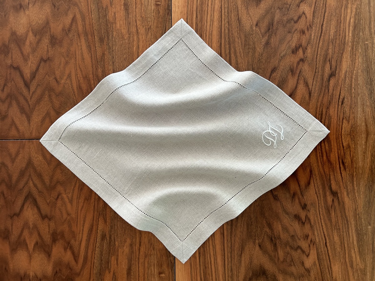 Ein quadratisches Leinen-Serviette in Naturfarbe mit Monogramm-Detail auf einer hölzernen Tischplatte.