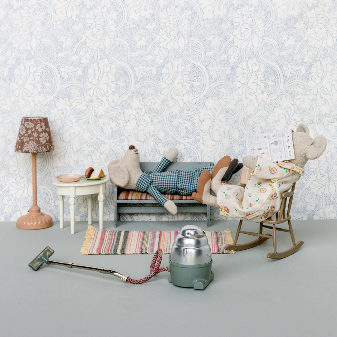 Zwei Spielzeugmäuse im Wohnzimmer, Oma und Opa Maus, die eine umfasst eine Zeitung in einem Schaukelstuhl, die andere liest auf einem Sofa. Umgeben von Miniaturmöbeln wie ein Tisch, eine Lampe, ein Staubsauger und ein Teppich.
