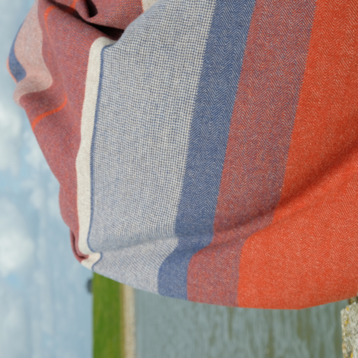Eine Decke mit Streifenmuster in den Farben rot, blau, grau und weiß. Die Decke ist aus Baumwolle gefertigt und liegt ausgebreitet vor einem Hintergrund mit Himmel und Graslandschaft.