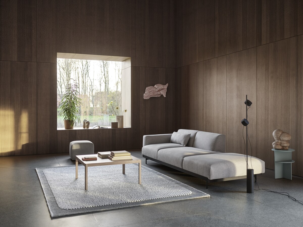 Modernes Wohnzimmer mit einer grauen Couch, einem Couchtisch aus Holz, einem grauen Teppich und einer schwarzen Bodenlampe »Post«. Im Hintergrund ein großes Fenster mit Pflanzen, das viel Tageslicht hereinlässt. Die Wände sind holzvertäfelt.