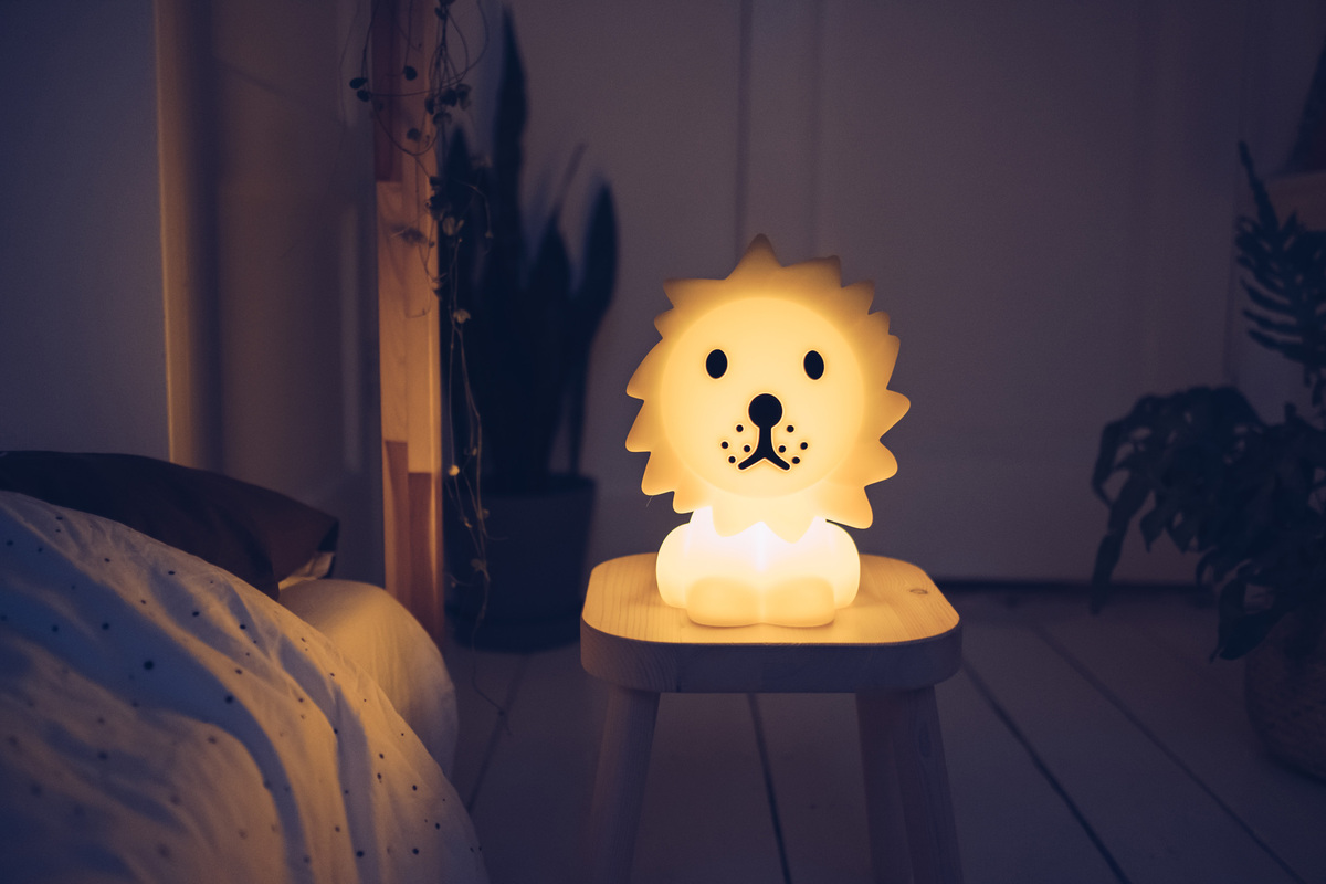 Ein Nachtlicht in Form eines niedlichen Löwen steht leuchtend auf einem Holzhocker in einem gemütlich eingerichteten Raum. Die warme Beleuchtung schafft eine beruhigende Atmosphäre vor dem Hintergrund eines Bettes und einigen Zimmerpflanzen.
