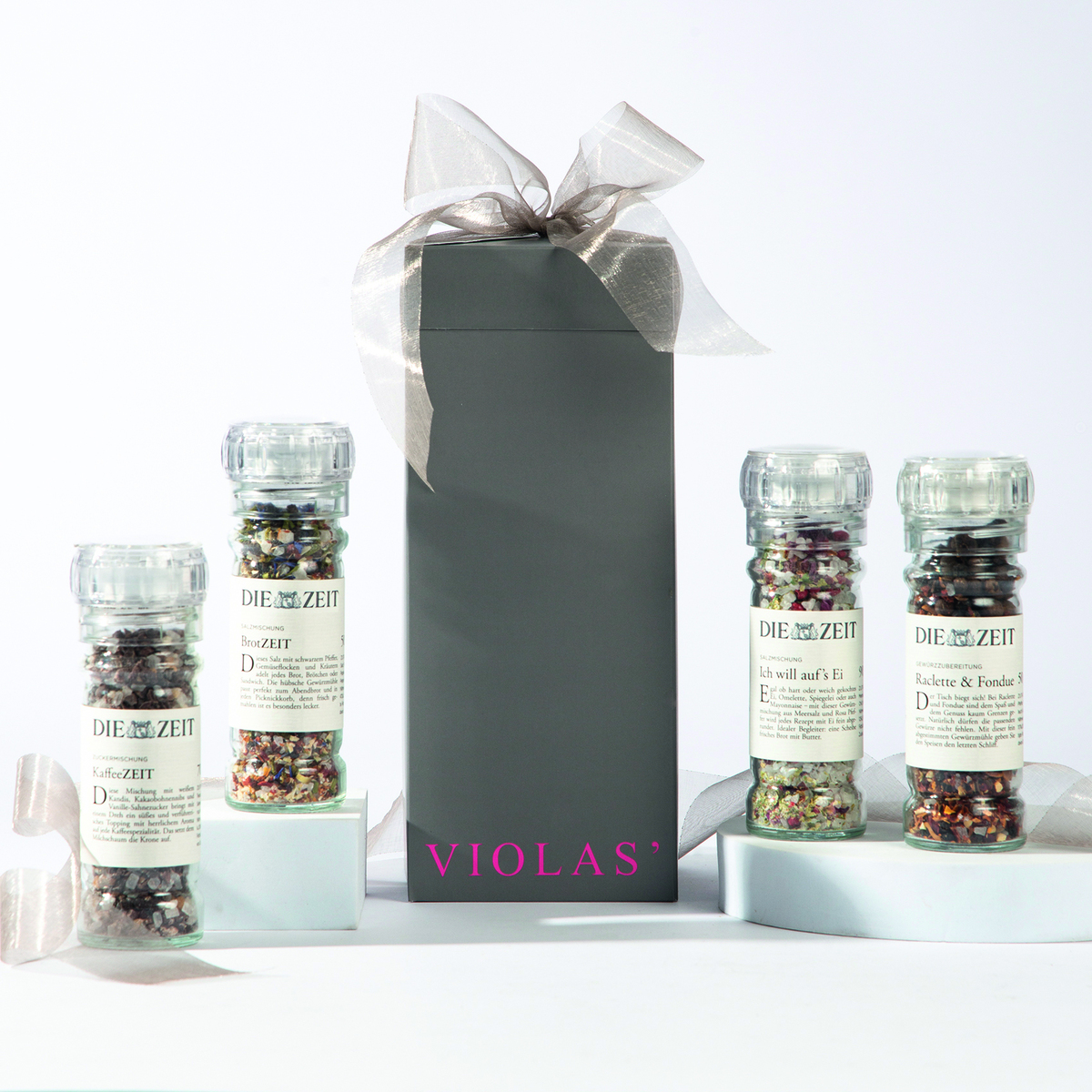 Das Bild zeigt ein 4er Gewürzmühlen-Set von ZEIT in einer eleganten Geschenkverpackung. Die verpackung ist schwarz mit einer edlen Schleife verziert und die Gewürzmühlen enthalten unterschiedliche farbenfrohe Gewürzmischungen. Auf der Verpackung ist der violette Schriftzug 'Violas*' zu sehen.