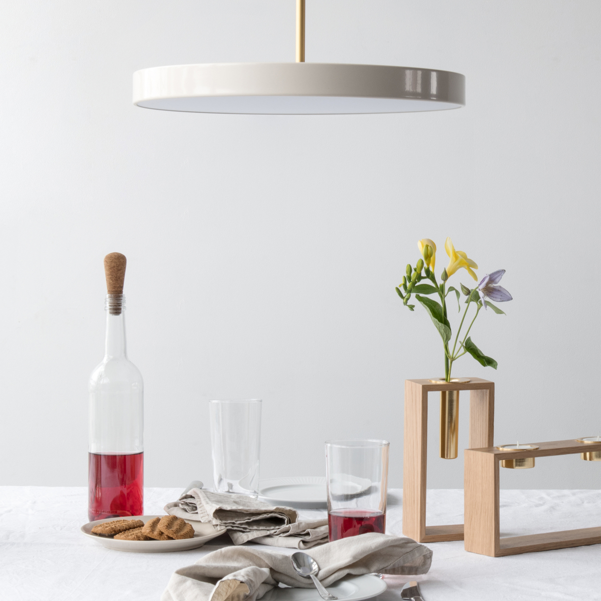 Elegante Pendelleuchte »Asteria Medium« über einem Esstisch, der mit einer Tischdecke, Gläsern, einer Flasche, Blumen und Besteck dekoriert ist.