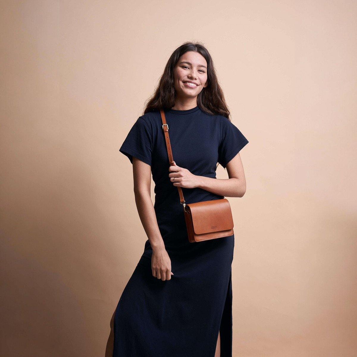 Eine junge Frau trägt ein dunkelblaues Kleid und hält eine kleine braune Ledertasche mit einem Schulterriemen, benannt 'Audrey Mini', vor einem beigen Hintergrund.