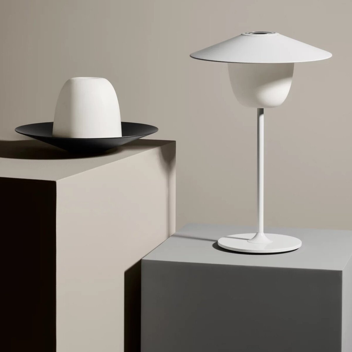 Eine mobile LED Tischleuchte namens ANI, die in einem minimalistischen, modernen Design gehalten ist. Die Lampe hat einen weißen Lampenschirm und einen schlanken, eleganten Fuß. Ein weiteres Modell hängt von der Decke, während ein kleinerer, zylinderförmiger Lampenschirm auf einem niedrigeren Podest steht. Die Farben im Bild sind hauptsächlich Weiß und Grautöne.
