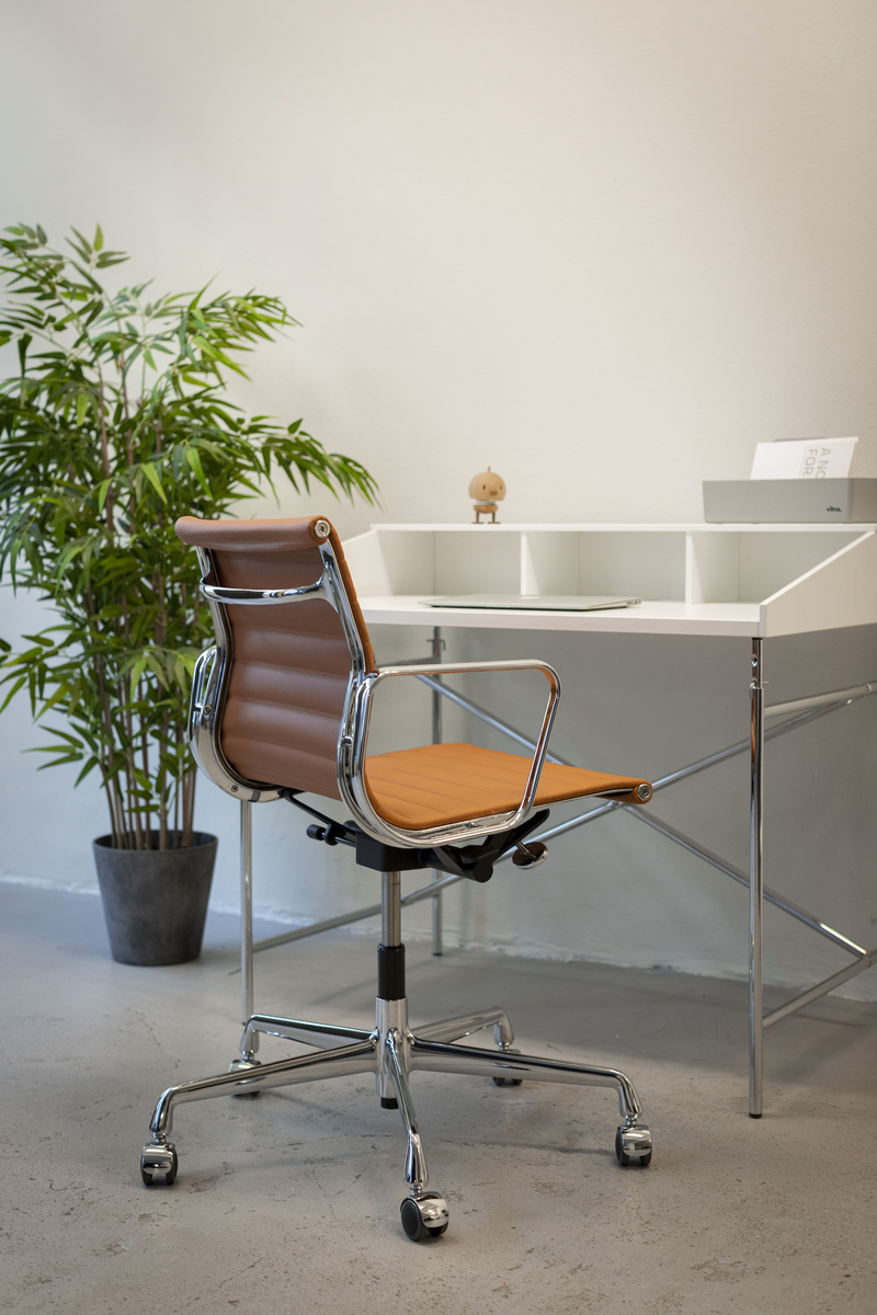Ein stilvoller Bürostuhl des Modells ZEIT-Sonderedition »Vitra Alu Chair EA 118« in verchromter Ausführung. Der Stuhl steht vor einem modernen weißen Schreibtisch mit mehreren Fächern. Im Hintergrund ist eine grüne Pflanze in einem grauen Topf zu sehen. Der Boden ist aus Beton und die Wände sind weiß.