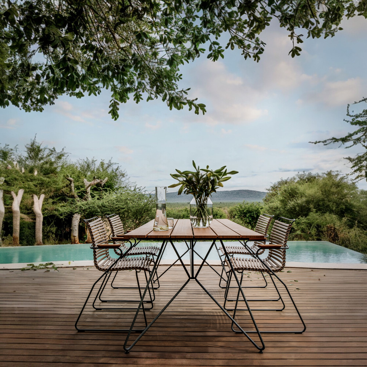 Ein moderner Gartentisch namens 'SKETCH' steht auf einer hölzernen Terrasse vor einem Pool mit üppiger Vegetation im Hintergrund. Der Tisch hat eine natürliche, braune Oberfläche mit einem schlanken, schwarzen Stahlrahmen. Einige passende Stühle aus Metall und Holz sind um den Tisch herum angeordnet. Die Szene vermittelt einen Eindruck von Ruhe und Naturgenuss.