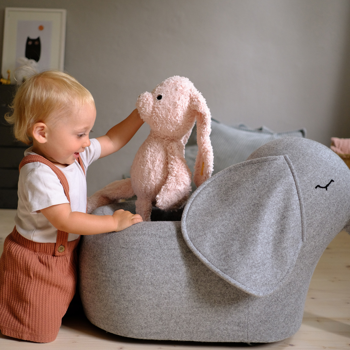Ein Kleinkind spielt mit einem weichen, grauen Elefanten-Spielzeug, das auf Rädern steht. Neben dem Elefanten befindet sich ein rosa Kuscheltier. Im Hintergrund ist ein Kinderzimmer zu sehen mit einem kleinen Regal.