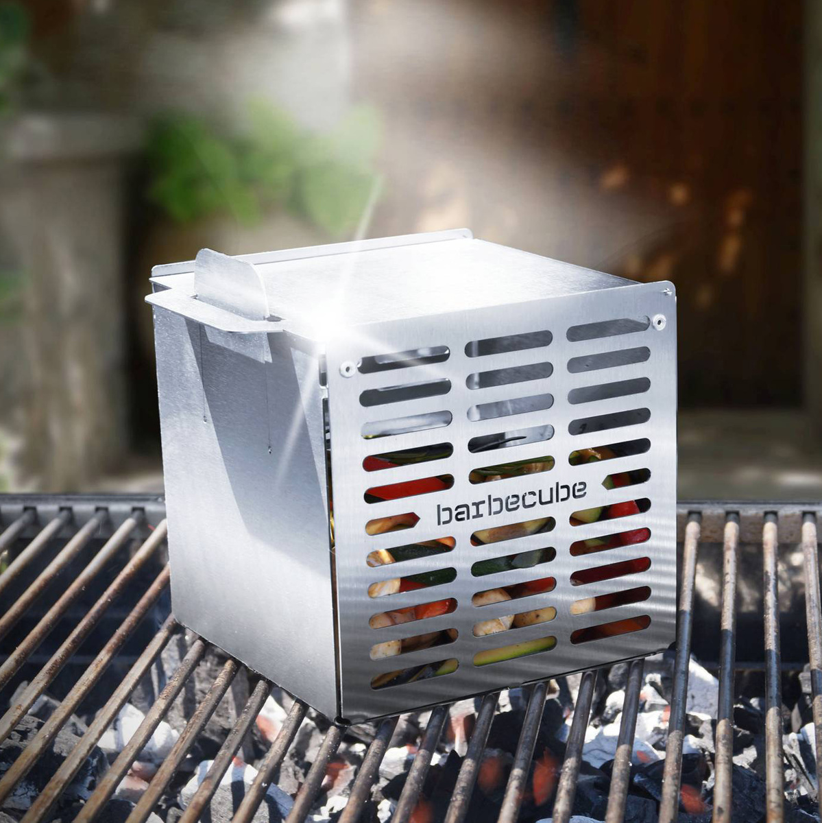 Ein silberner Grillwürfel namens 'The Cube' ist auf einem Grillrost zu sehen. Im Hintergrund befinden sich verschwommene Gartenpflanzen und eine dunkle Holztür. Lichtstrahlen fallen auf den Grill, was den Raucheffekt betont.