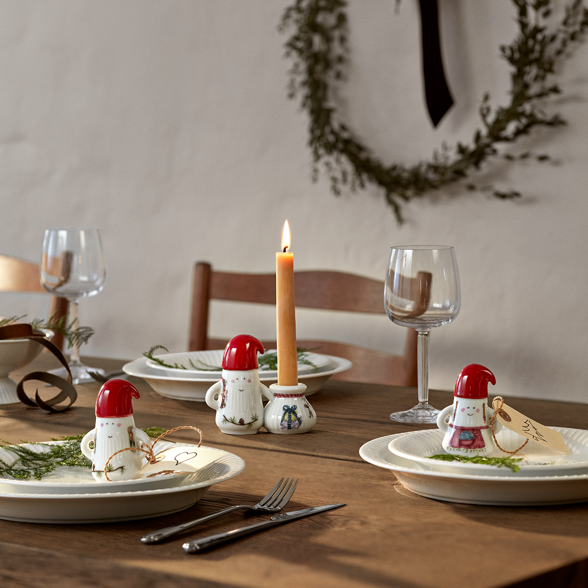 Eine festlich gedeckte Tafel mit Keramikfiguren in Form von Weihnachtsmännern der Serie »Hammershøi«, ergänzt durch einen Teller mit Tannenzweig, einem Weinglas und brennender Kerze. Die Szene vermittelt eine gemütliche, weihnachtliche Stimmung und eignet sich als Dekorations- oder Geschenkidee für die Advents- und Weihnachtszeit.