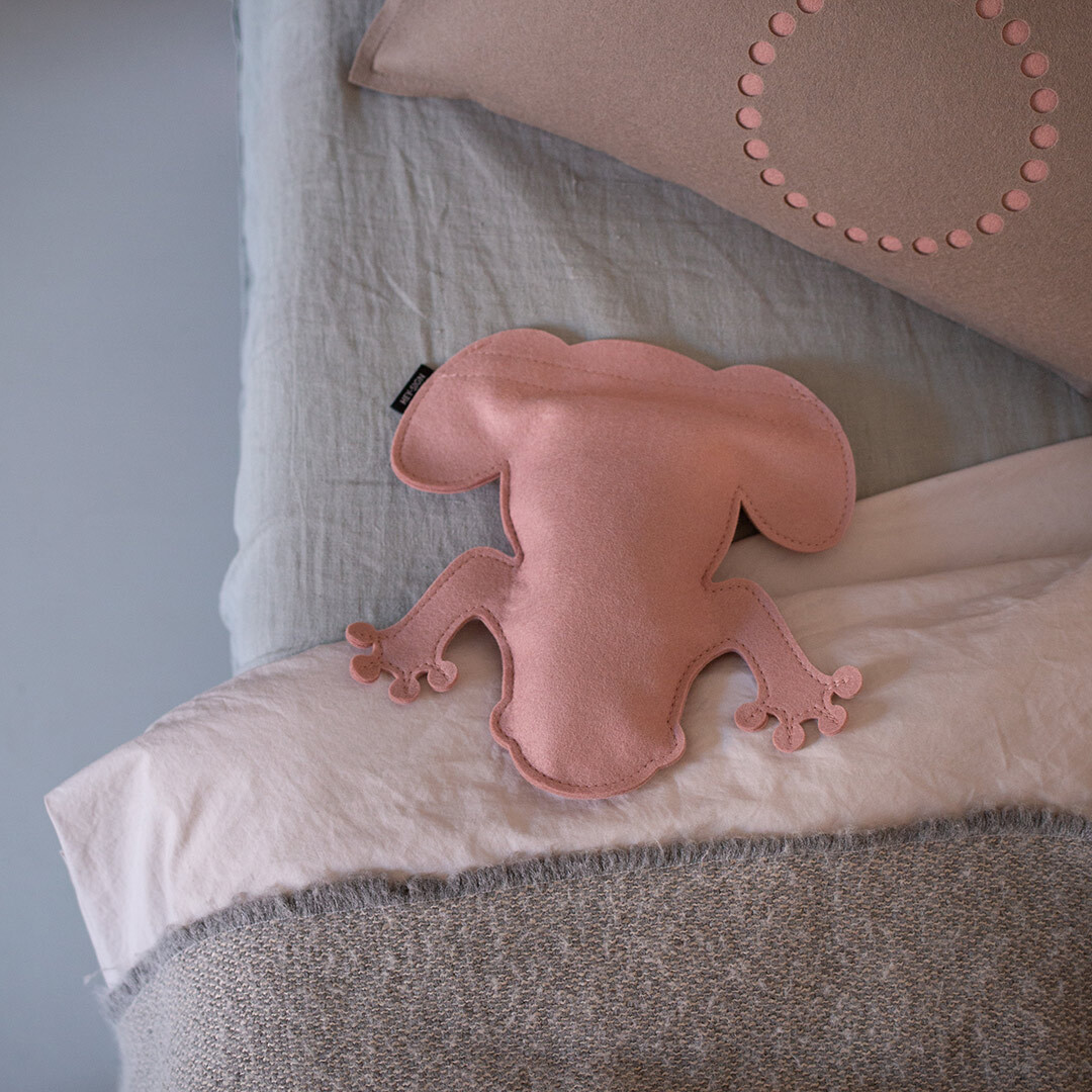 Auf dem Bild sitzt ein rosa Kirschkernkissen in Form eines Frosches namens »Frosch Nico« auf mehreren übereinandergelegten Kissen in Beige, Weiß, Grau und Blau.