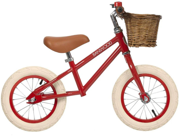 Ein rotes Retro-Laufrad für Kinder mit einem braunen Ledersitz und einem geflochtenen Holzkorb vorne. Die Reifen sind weiß, und das Design ist klassisch und ansprechend für Kinder.