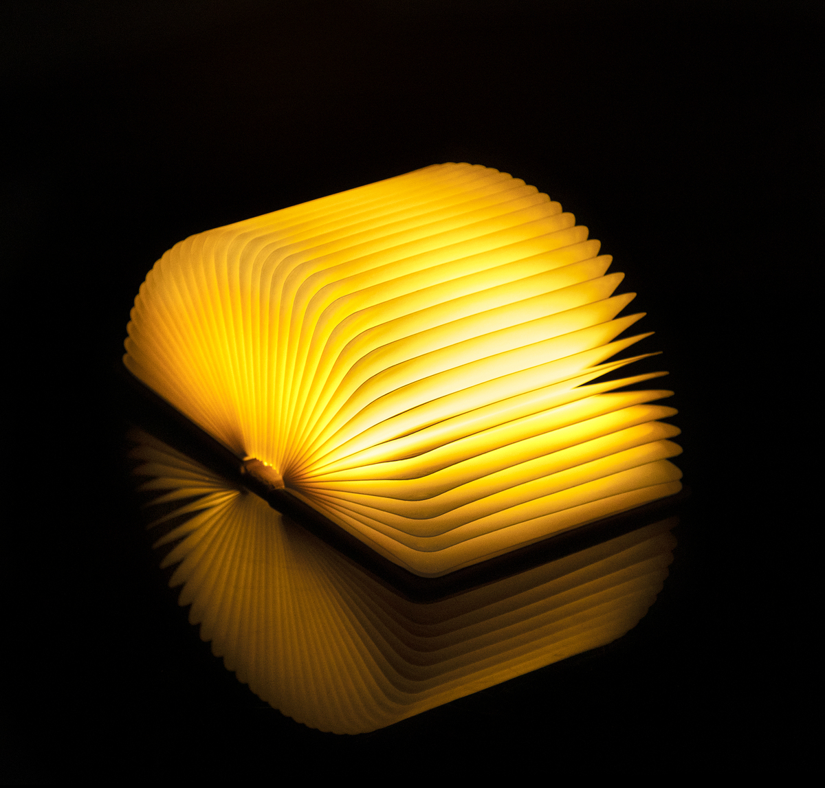 Ein aufklappbares Smart Booklight »Walnuss«, das in gelbem LED-Licht leuchtet. Es steht auf einem schwarzen Hintergrund und reflektiert sich selbst.