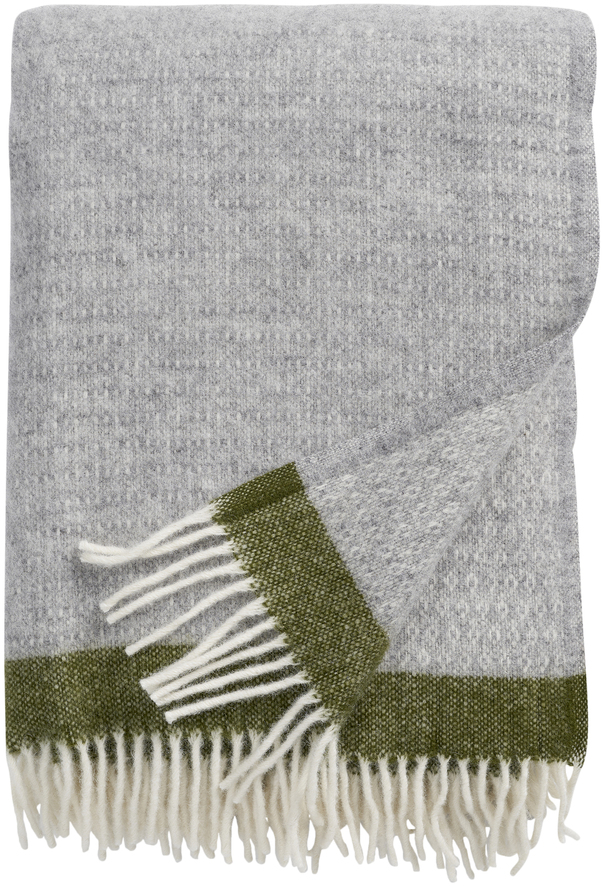 Ein großes, rechteckiges Plaid Hampus in Grau mit weißen und grünen Fransen liegt zusammengefaltet auf einem weißen Hintergrund. Das robuste Strickmuster und die gedeckten Farben passen gut in ein gemütliches Zuhause.
