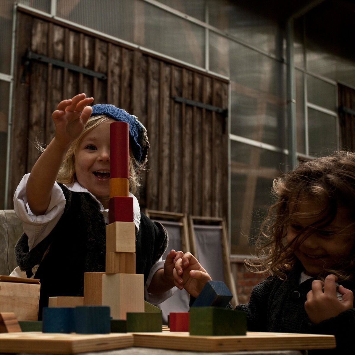 Zwei Kinder spielen mit Regenbogen Holzbausteinen. Das Spielzeug besteht aus bunten Holzklötzen in verschiedenen Farben und Formen. Im Hintergrund ist ein Raum mit großen Fenstern zu sehen.