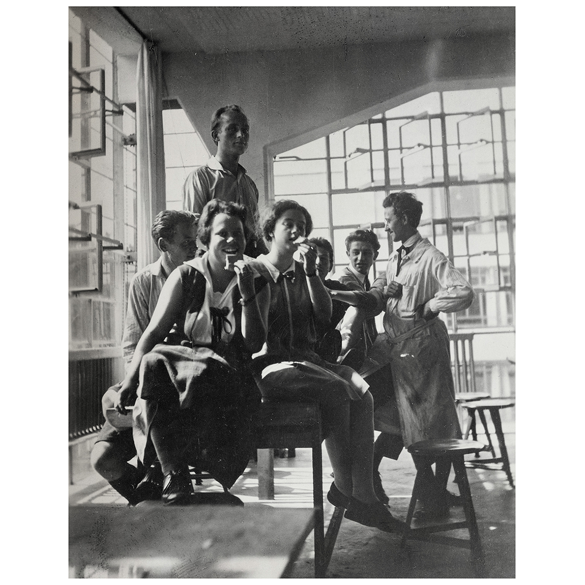 Historische Schwarzweißfotografie vom Bauhaus Archiv Berlin. Die Aufnahme zeigt eine Gruppe junger Menschen in einem lichtdurchfluteten Raum mit großen Fenstern und modernistischer Einrichtung. Das Bild illustriert die kreativen und gemeinschaftlichen Werte des Bauhauses in den 1920er Jahren.