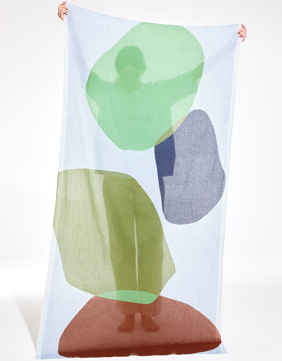 Großes Baumwolltuch mit transparentem, farbigem Muster in Grün, Blau, Braun und Grau. Eine Person hält das Tuch, wodurch ein interessantes Spiel von Farben und Licht entsteht.
