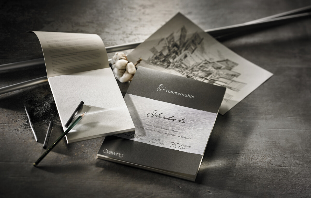 Ein Zeichenblock der Marke Hahnemühle namens 'The Collection »Sketch«', welcher 140-gsm-Skizzenpapier enthält. Im Bild liegt der Zeichenblock auf einem grauen Hintergrund und es sind einige künstlerische Werkzeuge sowie eine Skizze einer architektonischen Landschaft zu sehen.