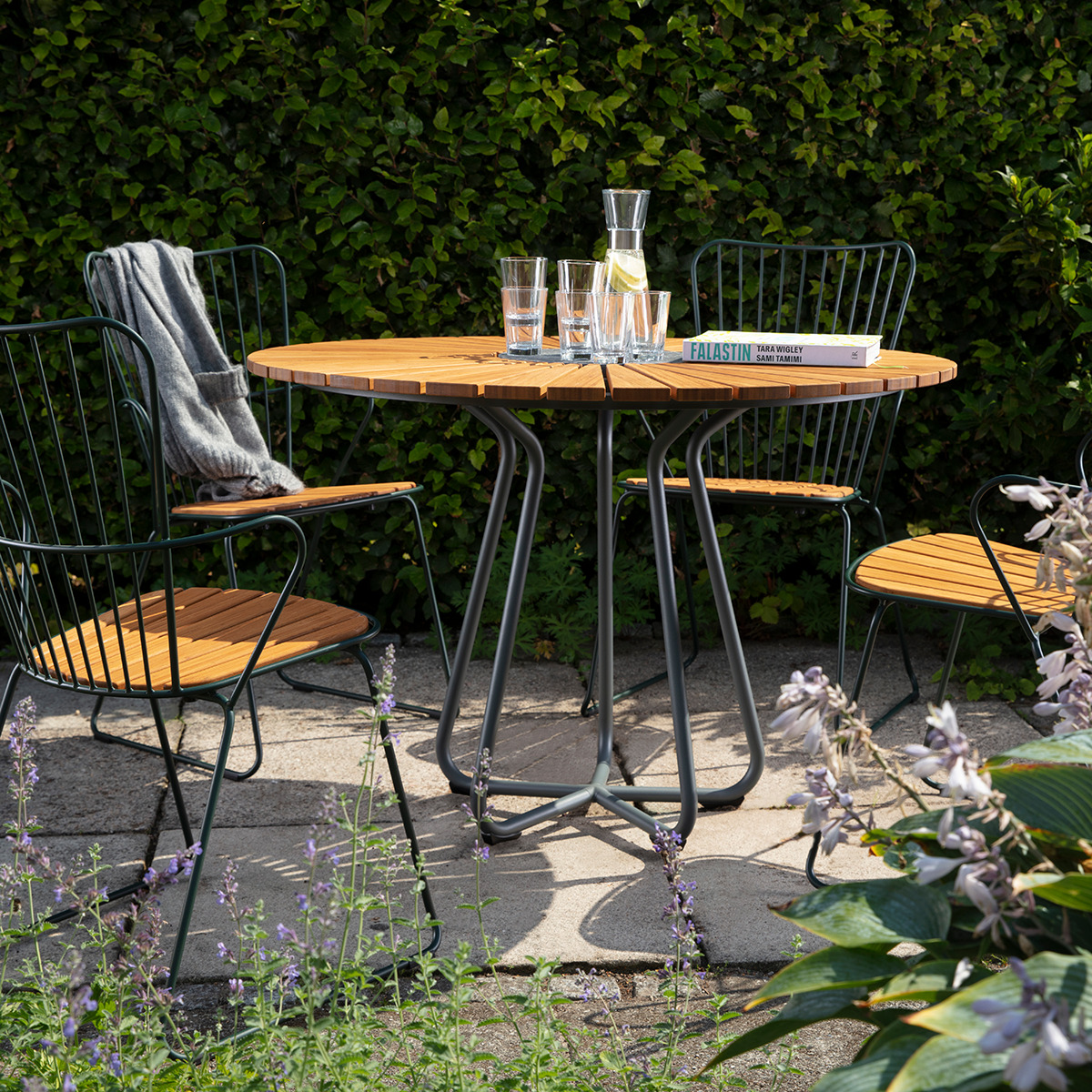 Runder Gartentisch »CIRCLE« aus Holz und Metall, umgeben von drei passenden Stühlen auf einer Terrasse mit grüner Hecke im Hintergrund. Auf dem Tisch stehen eine Karaffe, zwei Gläser und ein Buch. Sommerliches Gartenambiente mit Lavendelblüten im Vordergrund.