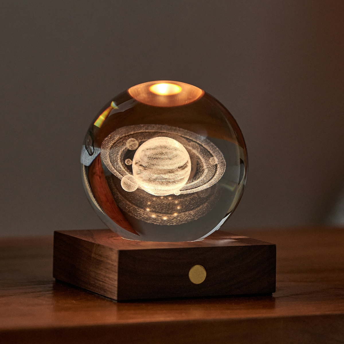 Eine dekorative Kugellampe namens »Amber Light« steht auf einem Holzboden. Sie leuchtet von oben in warmem Bernsteinlicht, in der Glaskugel ist ein 3D-Bild des Planeten Saturns eingraviert. Im Hintergrund befinden sich ein orangefarbenes Kissen und eine stilvoll platzierte Brille.