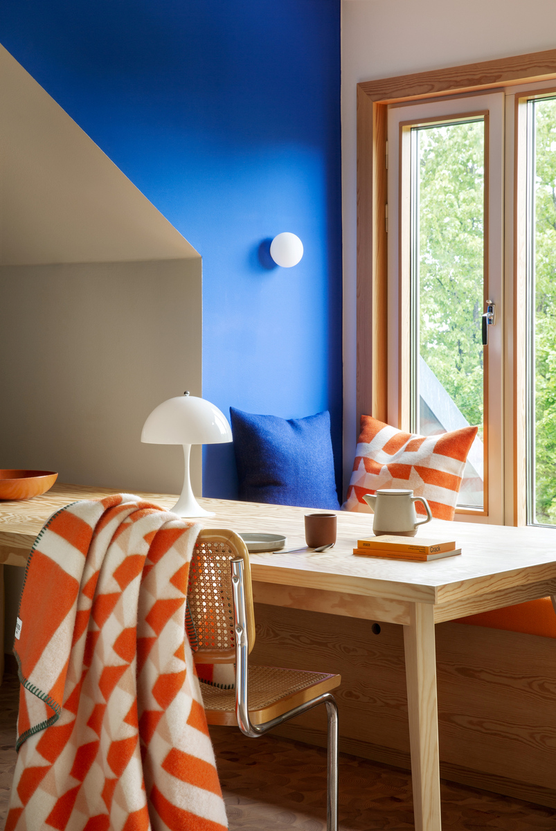 Ein heller Raum mit einem großen Holzfenster. Neben einem Holztisch steht ein Stuhl, auf dem eine orange-weiß gemusterte Lammwolldecke liegt. Im Hintergrund sind blaue Kissen und eine weiße Tischleuchte.