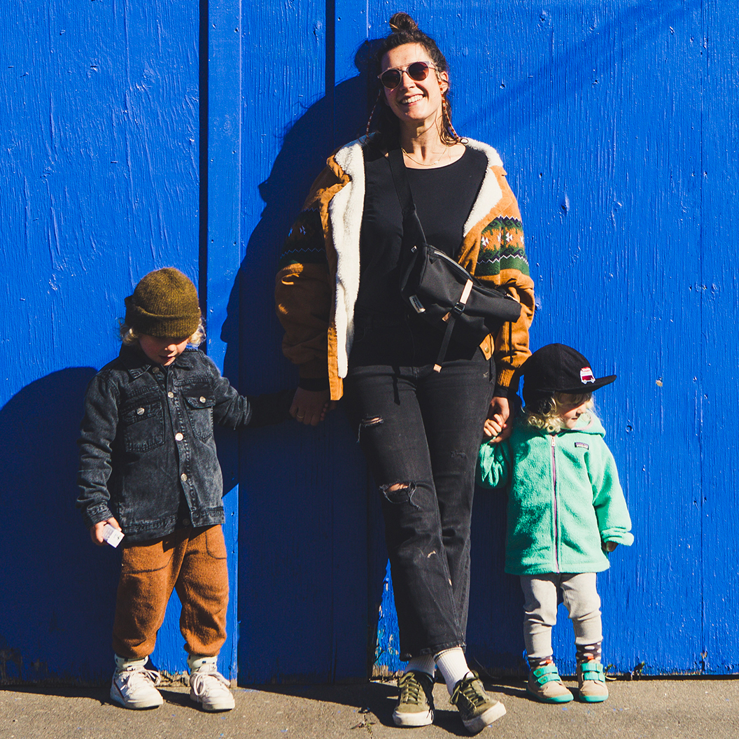 Eine Frau mit zwei Kindern steht vor einer blauen Wand. Sie trägt eine schwarze Tasche »Piha«. Die Frau ist modisch gekleidet und lächelt. Die Kinder tragen bunte Kleidung.
