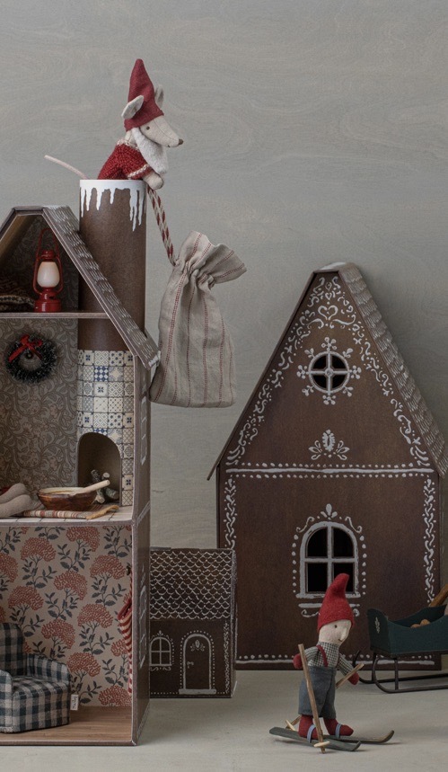 Eine Szene mit zwei niedlichen Weihnachtsmann-Mausfiguren. Eine Maus sitzt auf einem Holzhaus und hält eine gestreifte Geschenktüte, während die andere auf Skiern steht. Die Häuser sind dekoriert, eines davon wie ein Lebkuchenhaus bemalt, und die Figuren sind in Rot und Weiß gekleidet. Die Szene ist festlich dekoriert und erinnert an ein gemütliches Weihnachtsmärchen.