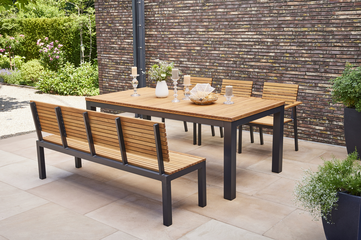 Ein moderner Esstisch aus Holz und Metall mit passender Gartenbank und Stühlen, platziert auf einer Terrasse mit natürlicher Gartenumgebung im Hintergrund.