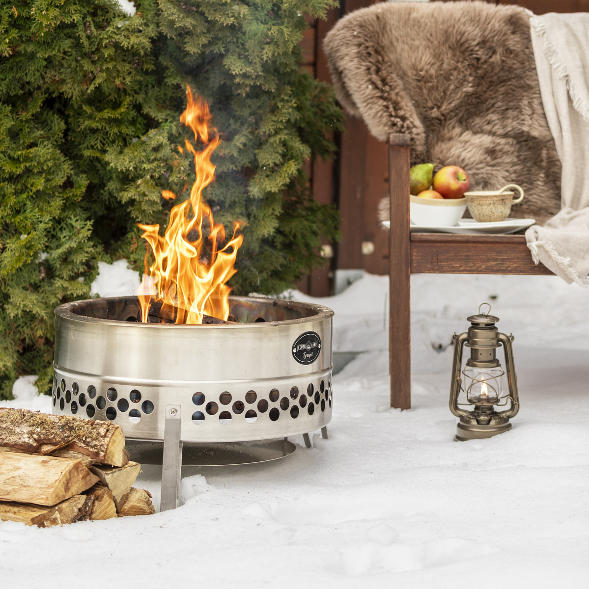 Eine Feuerschale namens »Tyropit« brennt lebhaft im verschneiten Garten. Im Hintergrund stehen Holzscheite, eine Laterne und ein gemütlicher Sitzbereich mit Decken und Kissen. Eine grüne Hecke und eine hölzerne Wand rahmen die Szenerie ein.