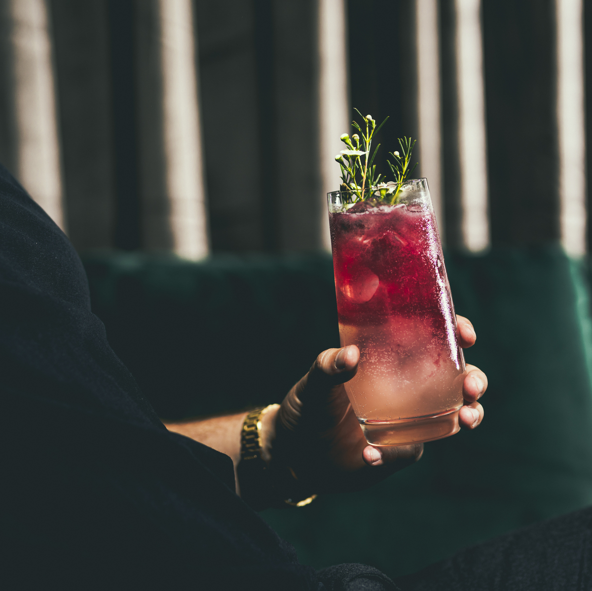 Ein Mann hält ein Glas mit einem gemischten Getränk, das mit Kräutern und Beeren verziert ist. Der Hintergrund ist dunkel mit einem Hauch von Grün.