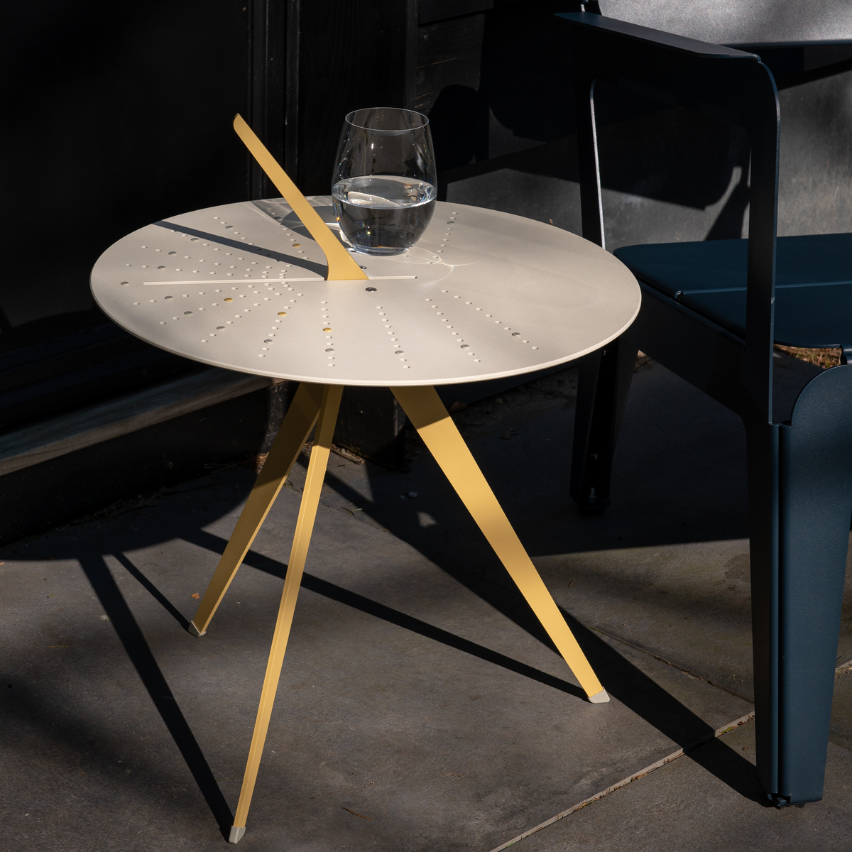 Moderner Tisch »Sundial« mit runder, beiger Tischplatte und gelben Beinen, draußen neben einem dunkelgrauen Stuhl auf einer Steinfläche. Ein Glas Wasser steht darauf im Sonnenlicht.