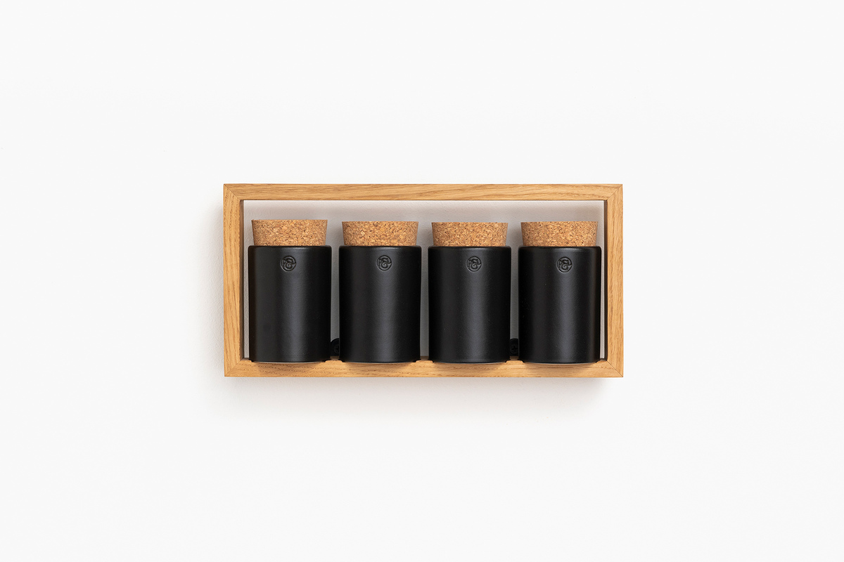 Das Bild zeigt ein Gewürzregal »bocuse«. Es besteht aus einem Holzrahmen mit vier schwarzen Behältern, die jeweils mit einem Korkdeckel verschlossen sind. Das Design ist modern und minimalistisch. Das Regal ist zur Wandmontage geeignet.
