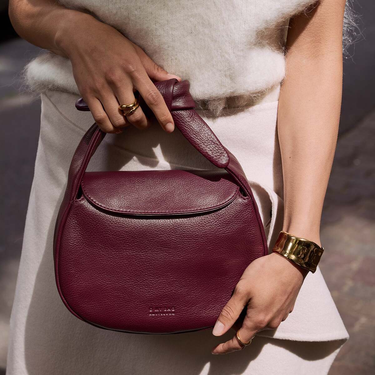 Eine Frau hält eine bordeauxrote Lederhandtasche mit elegantem Henkel. Sie trägt ein cremefarbenes, ärmelloses Oberteil und einen goldenen Armreif. Im Hintergrund ist ein unscharfer Außenbereich zu sehen.